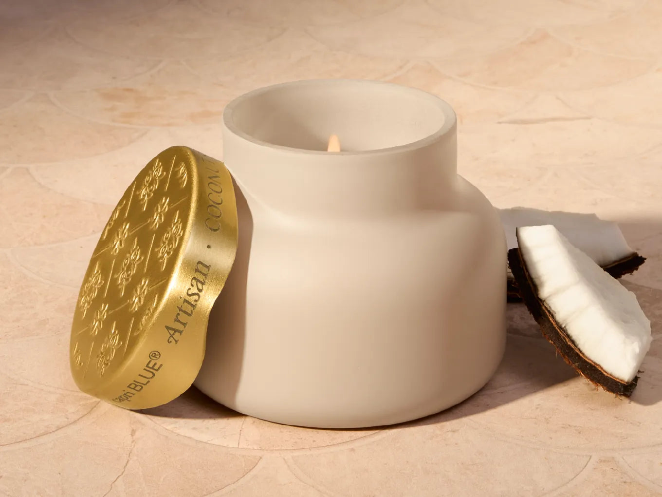 A matte off-white ceramic candle jar with a gold lid leaning against it, set on a beige stone surface next to a halved coconut.