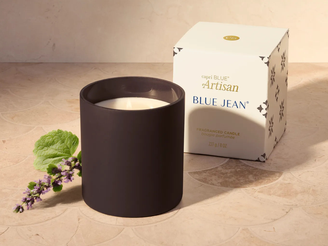 A matte dark brown candle jar with white wax sits beside its white branded box on a beige stone surface, with a small sprig of lavender and green leaves as styling props.