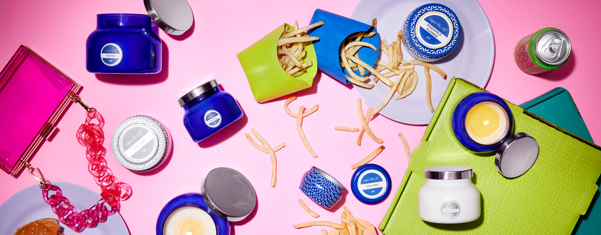 Various candles on a pink surface surrounded by french fries, a purse, soda can and pizza box