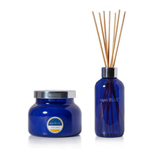 A blue glass reed diffuser set with a matching jar and several natural wood reeds, on a clean white background. The jar features a silver lid and a circular label that reads 'capri BLUE' and 'VOLCANO'.