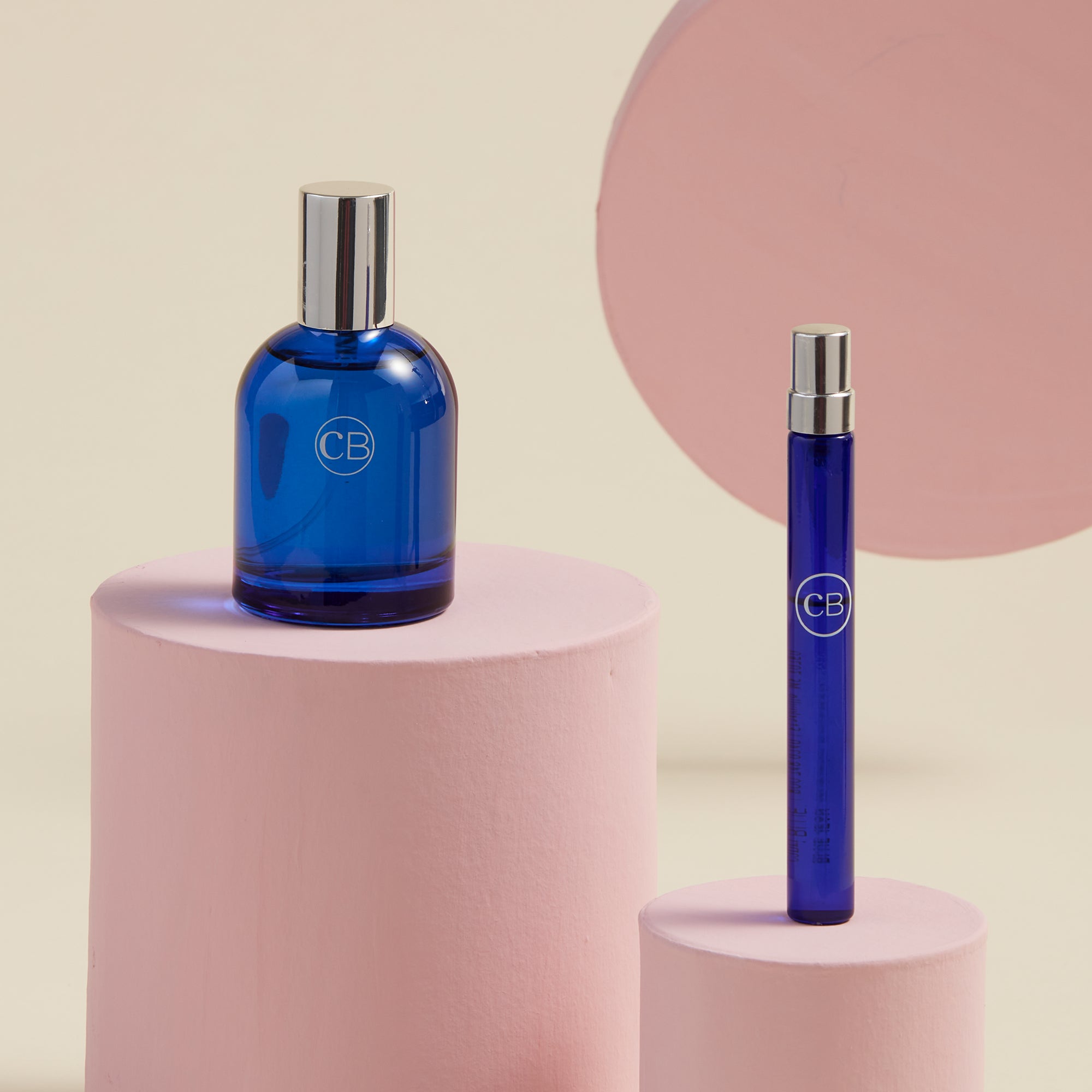 Two blue perfume bottles with silver caps on pink cylindrical stands against a beige background