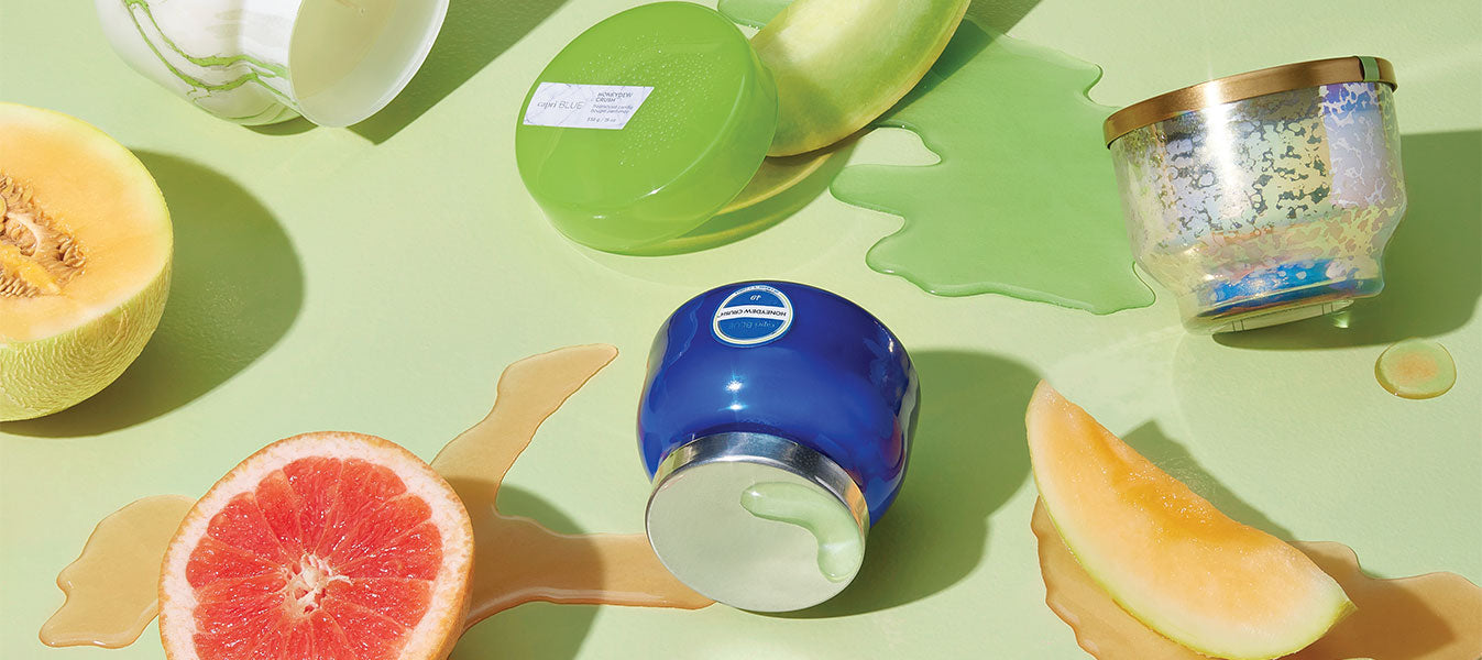 Various Honeydew Crush candles on a green surface next to sliced melon and grapfruit