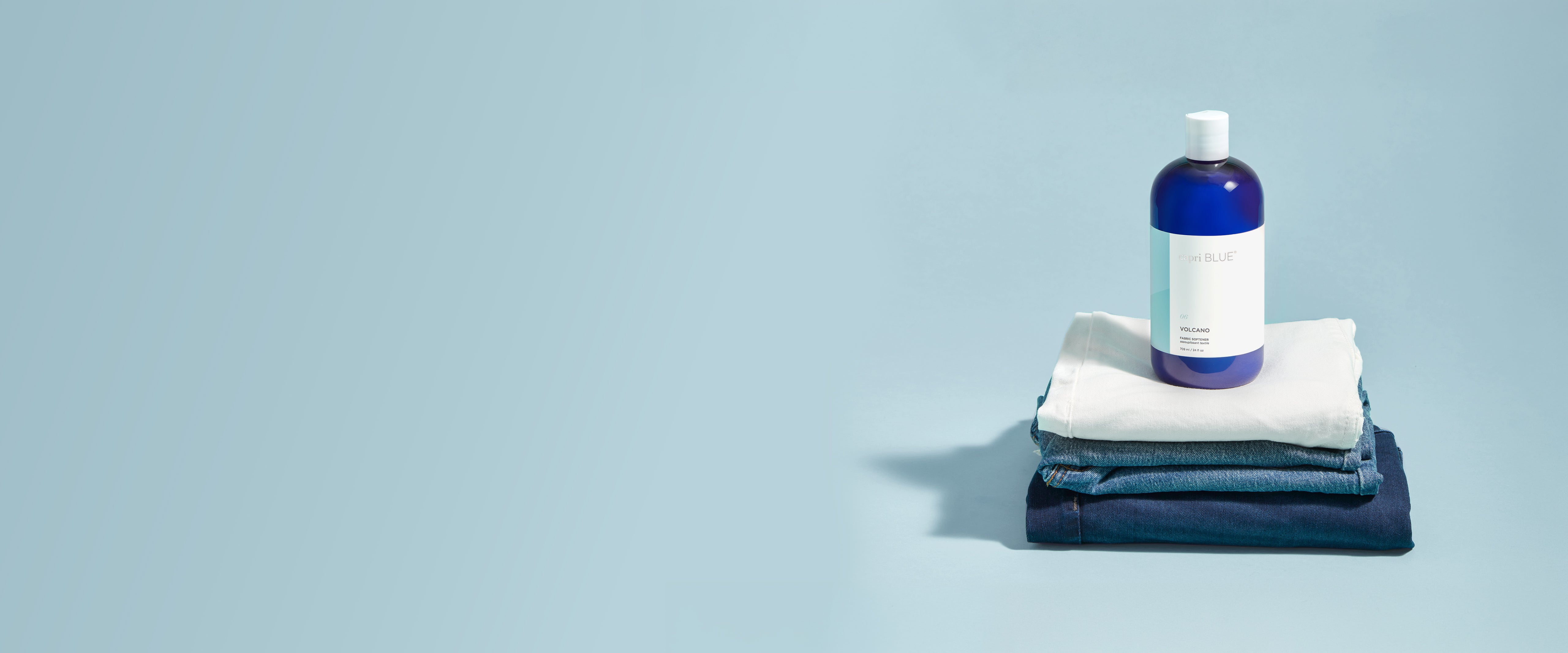 A blue bottle with a white cap and white label sits on top of folded white and dark blue towels against a light blue background.