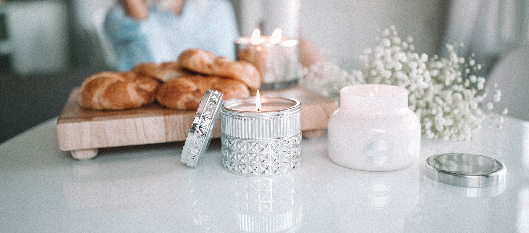 Two lit candles on a white surface with croissants on a cutting board in the background and flowers
