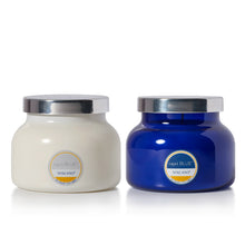 Two Volcano candles by Capri Blue on a white background, one in a cream-colored jar and the other in a cobalt blue glass jar, both with silver lids.